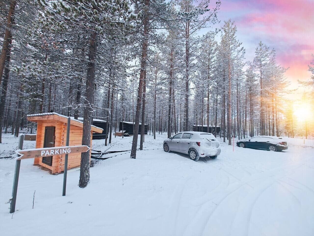 Lumipeitteinen metsä, jossa on puinen mökki, jonka kyltissä lukee ”PYSÄKÖINTI”, ja sen lähellä on parkkeerattuina kaksi autoa.