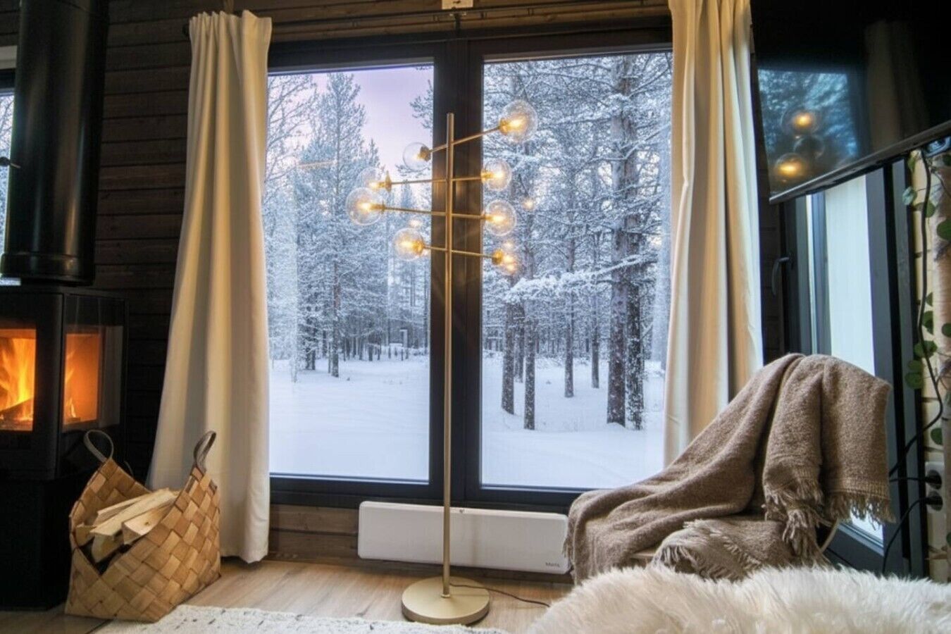 A cozy room with a fireplace, a large window with a view of a snowy forest, a floor lamp, and a basket of firewood.