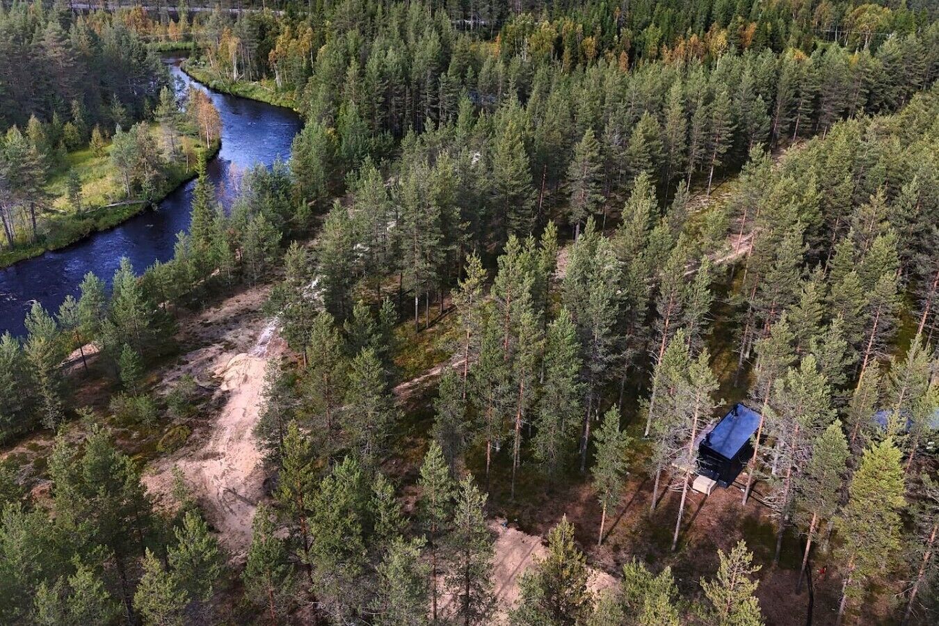 A forested area with a river, a dirt path, and a small building.