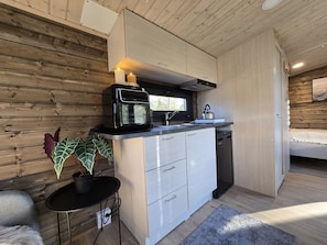 A compact kitchen with a microwave, coffee maker, and dishwasher. There is a potted plant on a small table and a bed in the adjacent room.