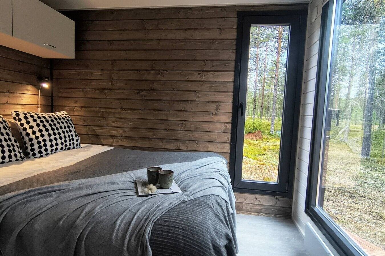 A bedroom with a large window, wooden walls, a bed with grey bedding, and a nightstand with two cups.