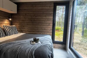 Un dormitorio con un ventanal grande, paredes de madera, una cama con ropa de cama gris y una mesita de noche con dos tazas.
