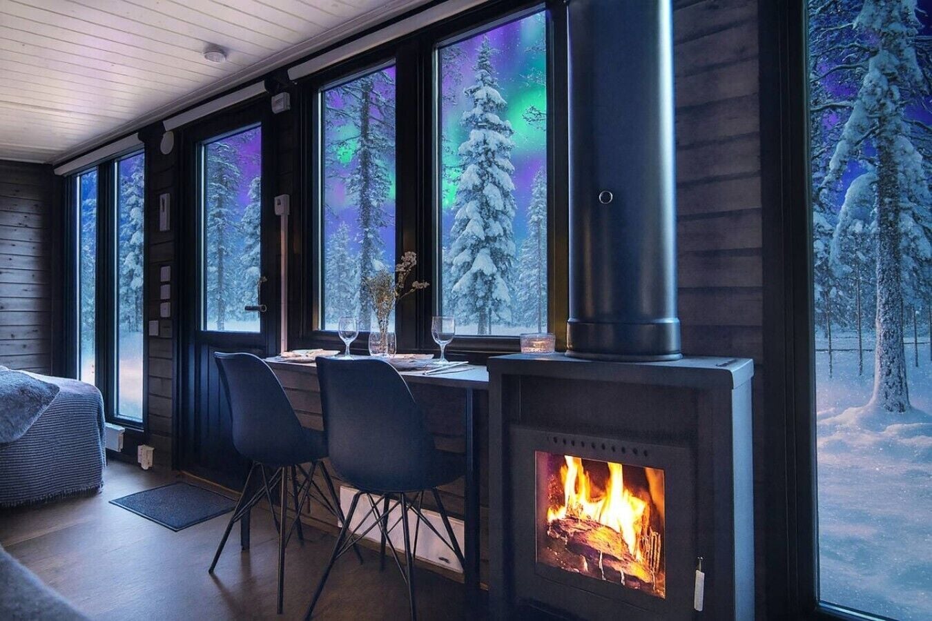 A cozy room with a fireplace, a dining table, chairs, and large windows with a view of snow-covered trees.