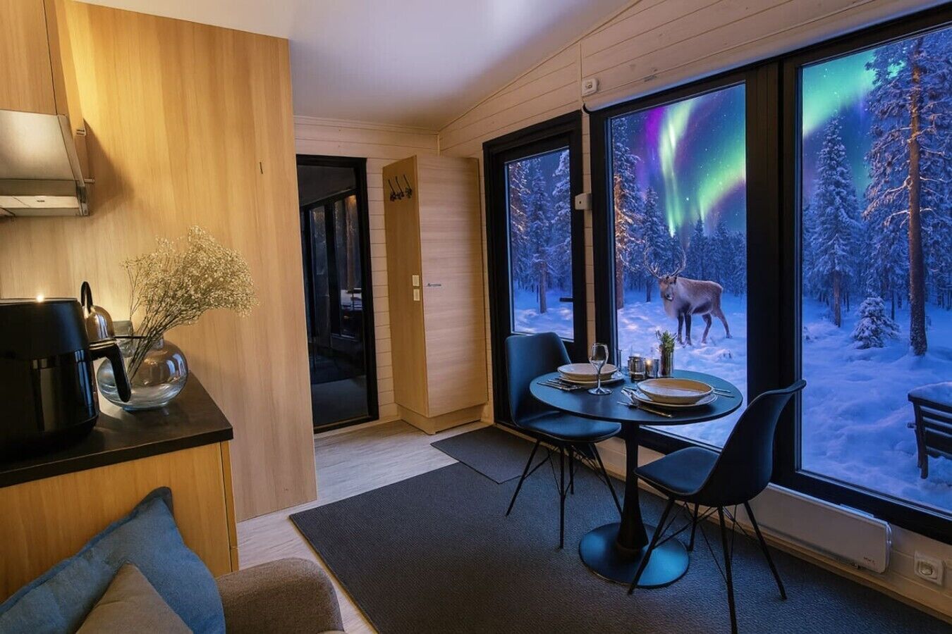 Un intérieur moderne comprenant un coin repas, un espace dédié à la préparation du café et de grandes fenêtres offrant une vue sur une forêt enneigée et des aurores boréales.