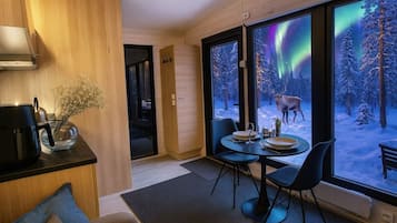 Et moderne interiør med spiseplads, kaffestation og store vinduer med udsigt over en sneklædt skov og nordlys.