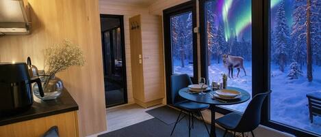 Et moderne interiør med spiseplads, kaffestation og store vinduer med udsigt over en sneklædt skov og nordlys.