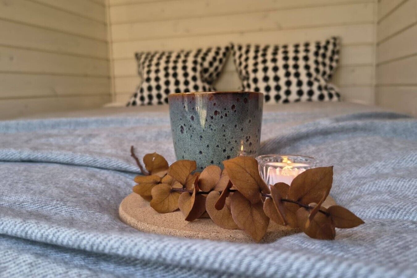 A bed with a textured blanket, a decorative candle holder with a candle, and dried leaves on a coaster.