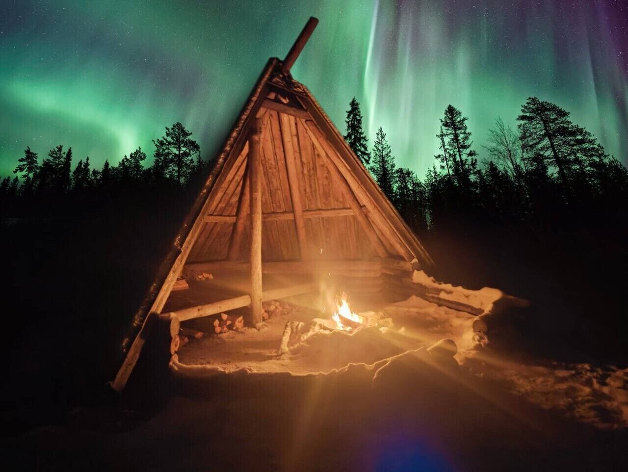 A wooden structure with a fire inside, surrounded by snow and trees, under a night sky with aurora borealis.