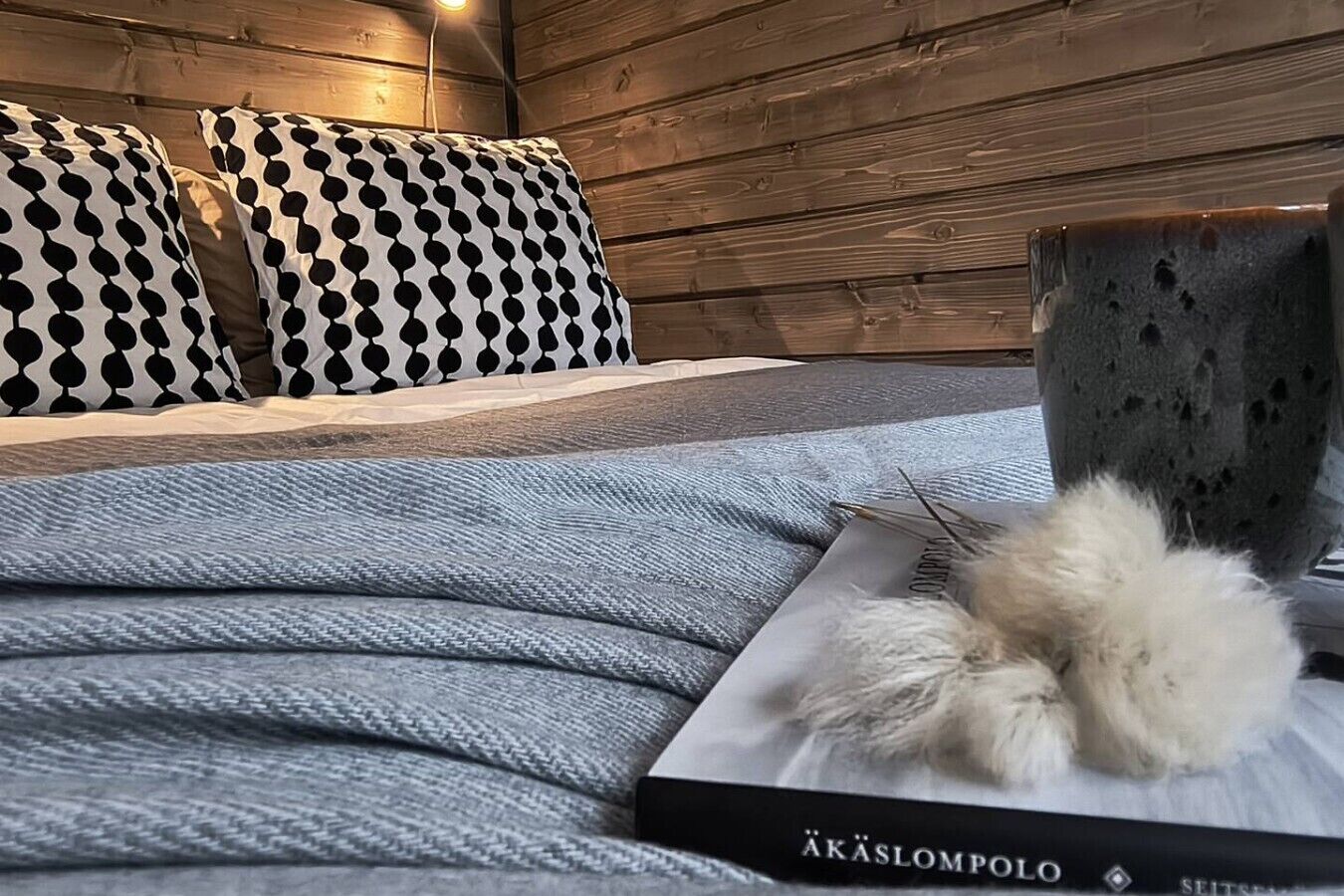 A bed with a patterned pillow, a grey blanket, and a book titled 'AKASLOMPOLO' on a wooden wall.