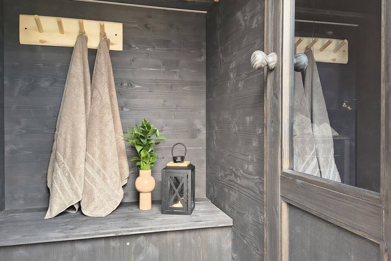 A dark wooden wall with a window, a towel hanging on a wooden rack, a potted plant, and a lantern on a ledge.