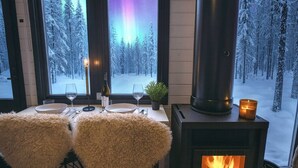 A cozy room with a fireplace, a dining table set for two, and a view of a snowy forest through large windows.
