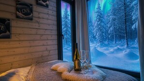 A cozy room with a wooden wall, a window with a view of a snowy forest, and a table with a bottle and glasses.