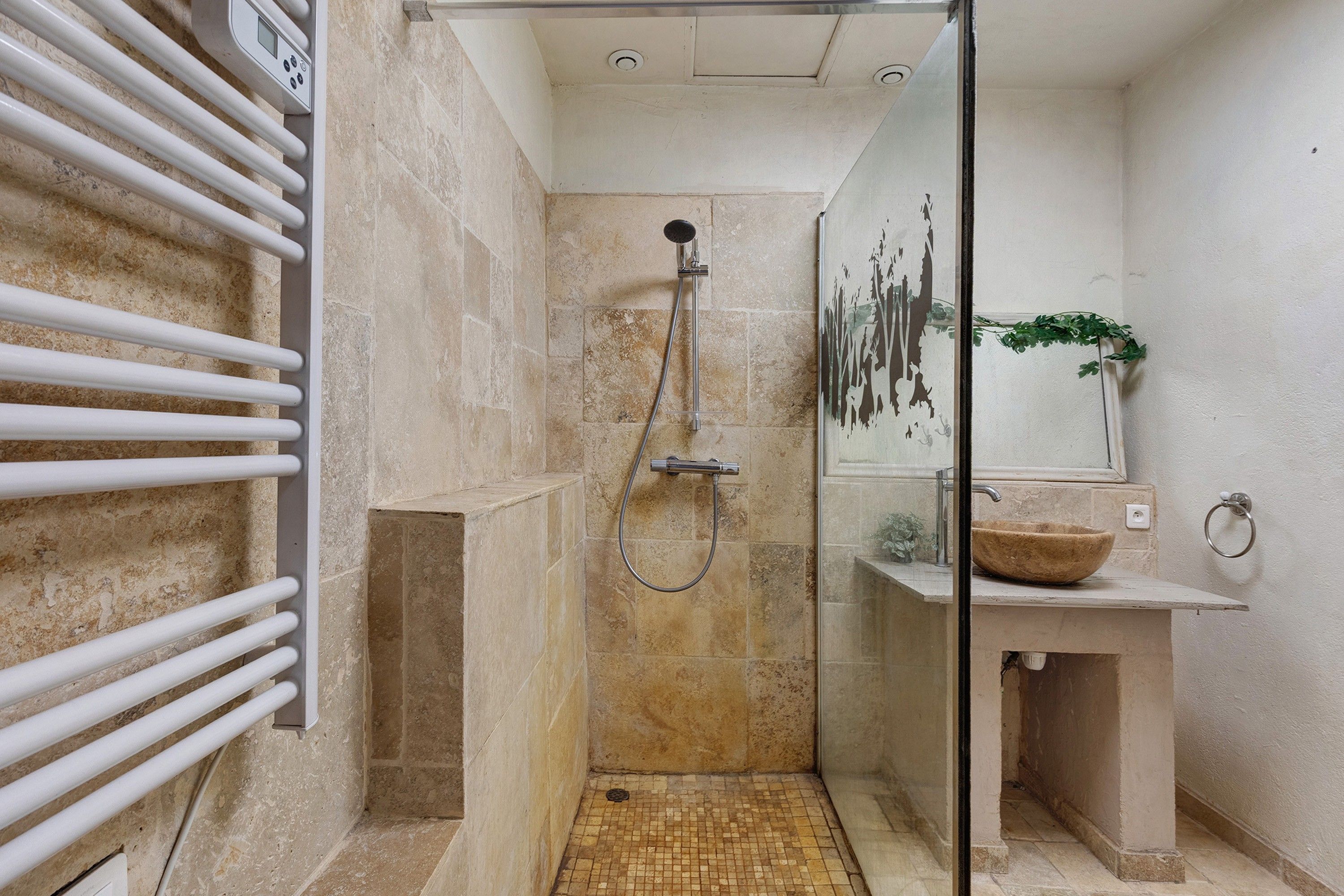 A glass-enclosed shower with a handheld showerhead, a white towel rack, and a stone-tiled floor.
