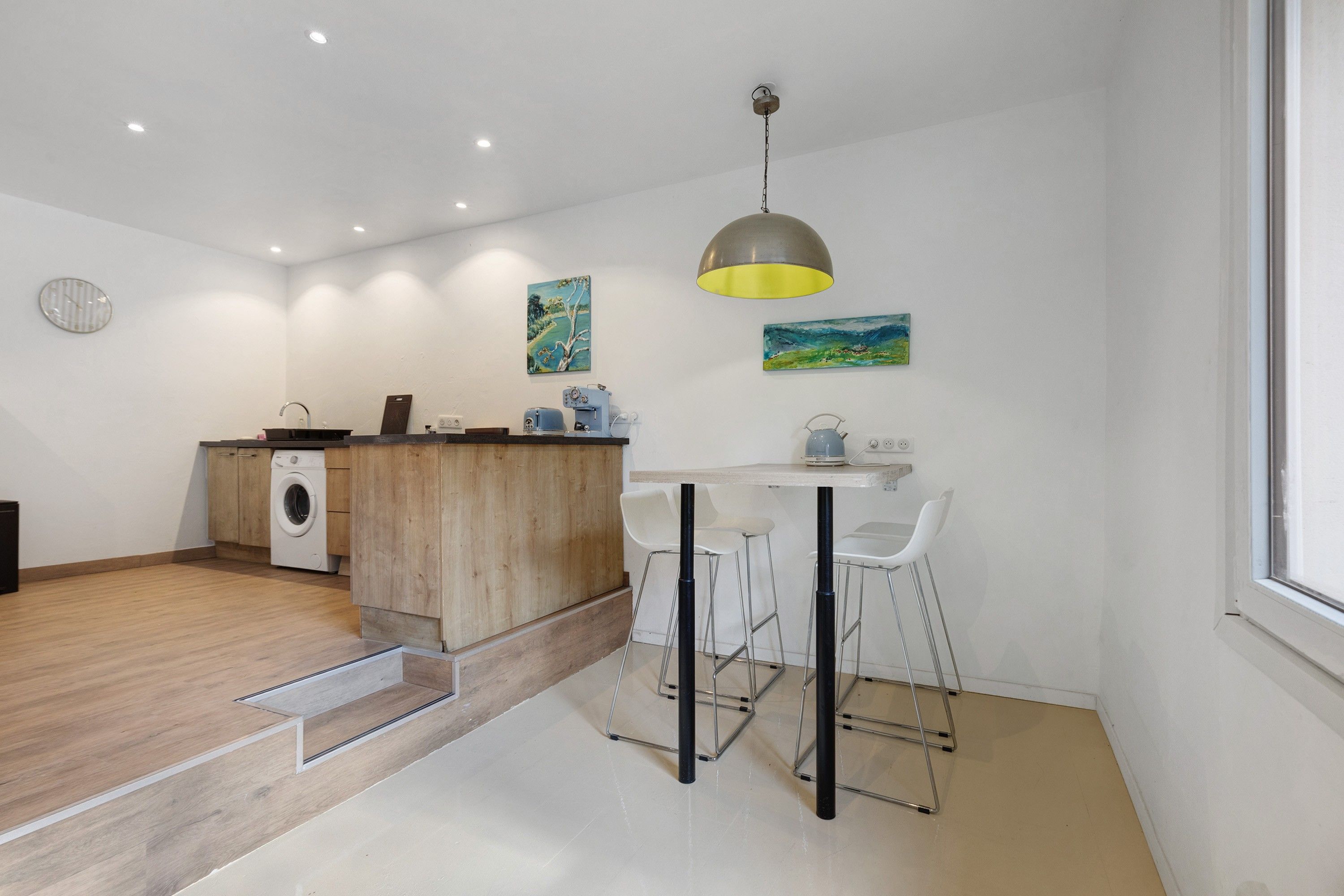 Cocina compacta con lavadora, encimera de madera y zona de comedor con taburetes de bar.