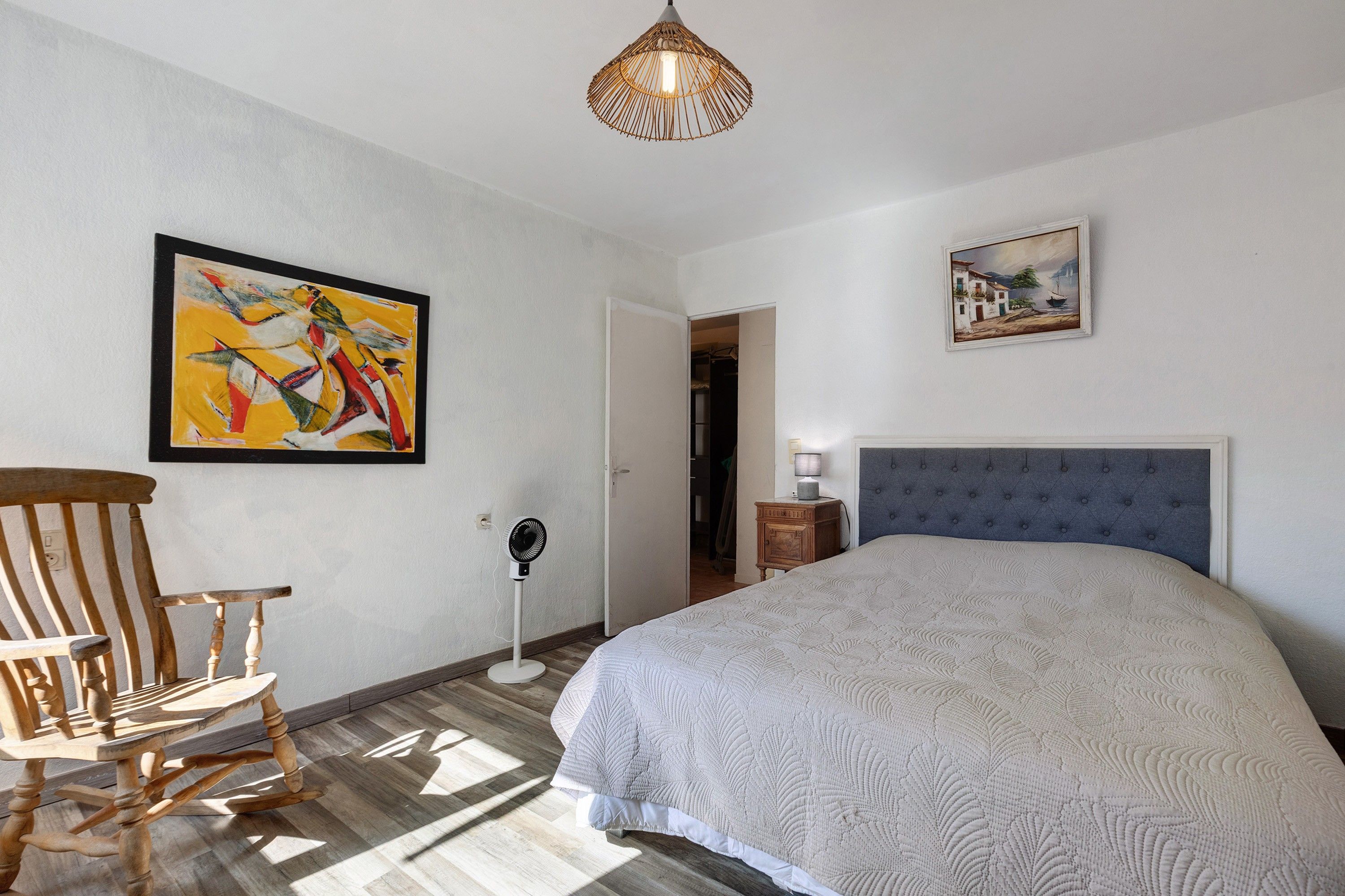 Ein Schlafzimmer mit einem Bett, einem Schaukelstuhl, einem Standventilator und Kunstwerken an den Wänden.