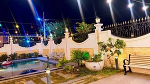 A nighttime outdoor pool area with a fountain, surrounded by a white wall with decorative elements, and a bench along the walkway.