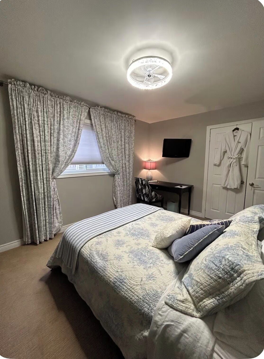 A bedroom with a bed, curtains, a lamp, a robe, and a television on the wall.