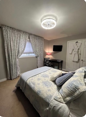 A bedroom with a bed, curtains, a lamp, a robe, and a television on the wall.