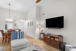 A living room with a flat-screen TV, a dining area with wooden chairs, a sofa, and a small cabinet with books. - Fairway Serenity at River Club (Pawleys Island)