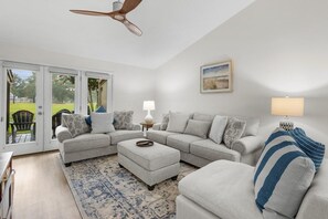 A living room with a sectional sofa, ottoman, and a ceiling fan. - Fairway Serenity at River Club (Pawleys Island)