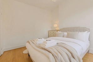 A neatly made bed with a fluffy white blanket, a bedside table with a lamp, and a nightstand with drawers. - Trendy 3BR Fulham Home Near Chelsea w/Garden (Greater London)