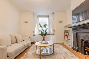 A living room with a sofa, a round coffee table, a plant, a fireplace, and a window with curtains. - Trendy 3BR Fulham Home Near Chelsea w/Garden (Greater London)