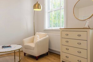 Una habitación con un sillón blanco, una mesita redonda pequeña, un aparador con cajones, una lámpara de pie y una ventana con persianas.