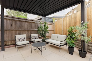 Une terrasse extérieure dotée d’un espace salon couvert, comprenant un canapé et des fauteuils, ainsi que deux plantes en pot.