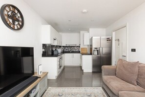 A modern kitchen with white cabinets, a large clock, and a flat-screen TV. - New! Cosy 2BR w/ Free Parking | Near Wimbledon (Saint Helier)