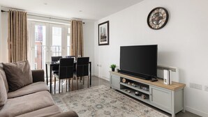 Una sala de estar con sofá, televisor de pantalla plana, comedor con sillas y alfombra estampada.