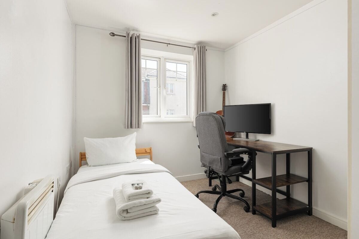 A bedroom with a bed, a desk with a computer, a guitar, and a window with curtains.