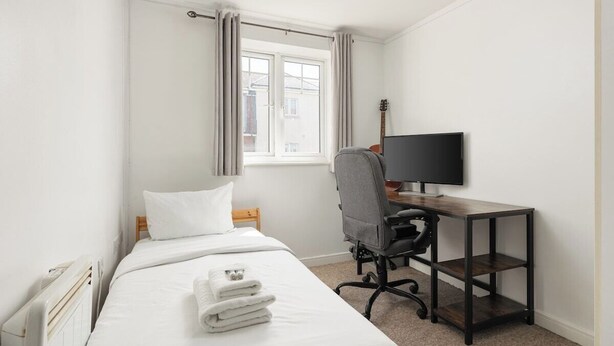 Un dormitorio con una cama, un escritorio con una computadora, una guitarra y una ventana con cortinas.