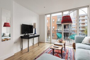 A modern living room with a flat-screen TV, a coffee table, and a sofa. There is a balcony with chairs and a red lamp. - New! Modern 3BR w/Balcony | Hoxton & Shoreditch (Greater London)