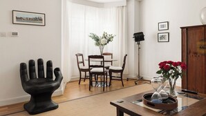A modern living room with a dining area, a black sculptural chair, a vase with red flowers, and a table with an hourglass. - New! Charming 2BR Flat in Chelsea (Greater London)