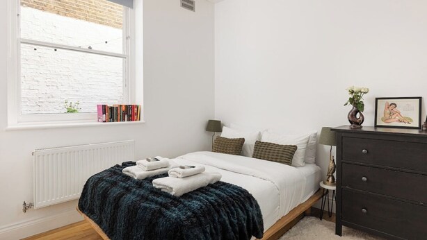 A bedroom with a bed, a dresser, a window with a view of a brick wall, and a radiator. - New! Charming 2BR Flat in Chelsea (Greater London)