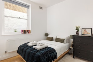 A bedroom with a bed, a dresser, a window with a view of a brick wall, and a radiator. - New! Charming 2BR Flat in Chelsea (Greater London)