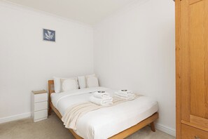A neatly made bed with white linens and towels, a small bedside table, and a wooden wardrobe. - New! Charming 2BR Garden Home | Hanger Lane (Greater London)