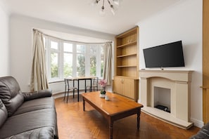 A living room with a sofa, a wooden coffee table, a fireplace, a television, and a bay window with curtains. - New! Charming 2BR Garden Home | Hanger Lane (Greater London)