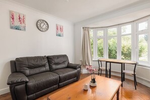 A living room with a leather sofa, a wooden coffee table, and a window with curtains. - New! Charming 2BR Garden Home | Hanger Lane (Greater London)