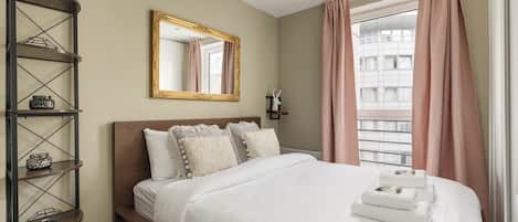 A hotel room with a bed, a mirror, a shelf with decorative items, and a window with curtains.