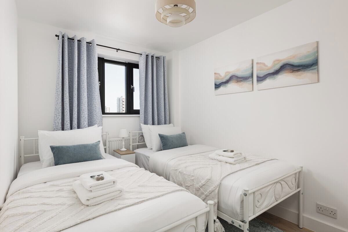 A bedroom with two beds, a window with curtains, a bedside table with a lamp, and abstract wall art.