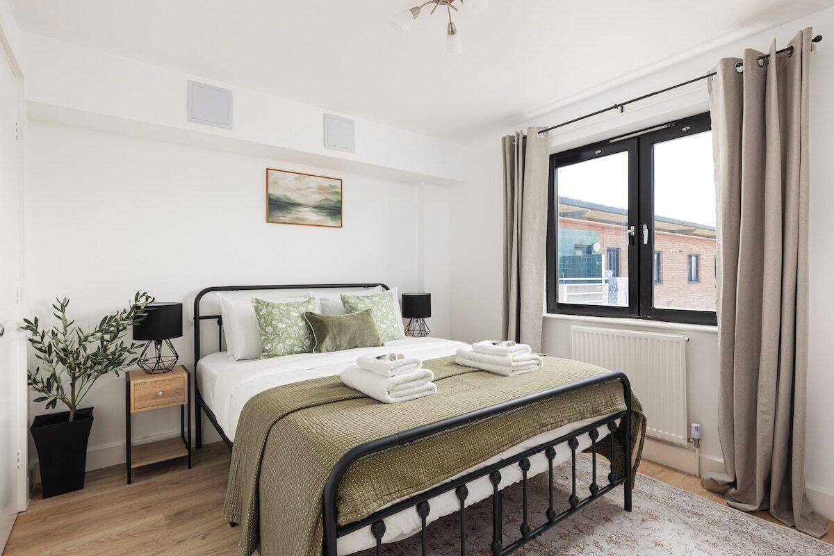 Ein Schlafzimmer mit Bett, Nachttischen, einem Fenster mit Vorhängen, einem Heizkörper und Blick auf Gebäude.