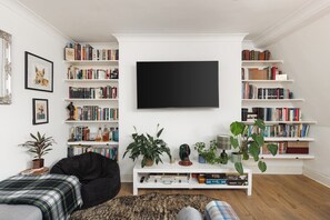 A living room with a large flat-screen TV, multiple bookshelves, a black bean bag chair, and a coffee table with decorative items. - New! Stylish Battersea 1BR | 6 Mins to Clapham Jct (Greater London)