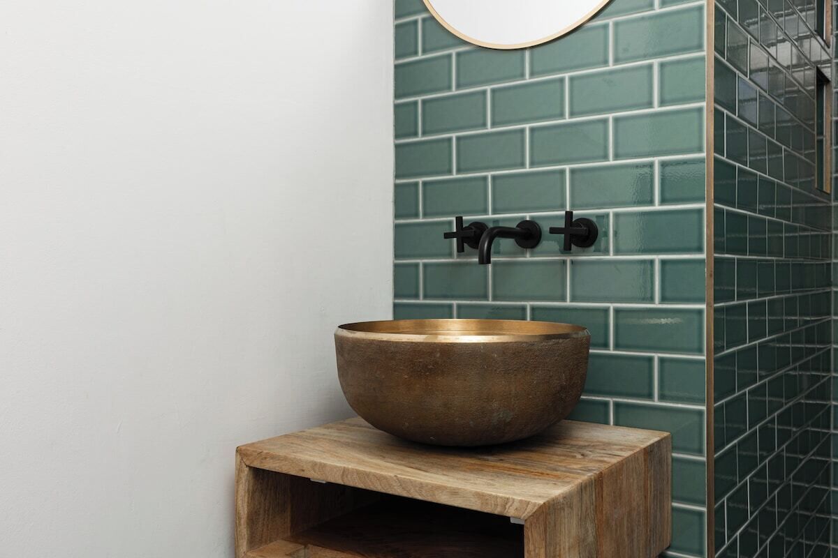 Un bagno con uno sgabello in legno, uno specchio rotondo e una parete rivestita di piastrelle verdi.