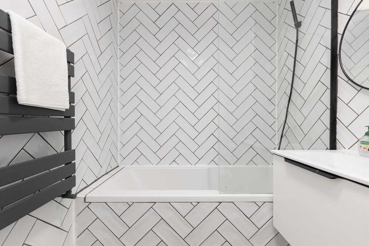 Une salle de bain avec un carrelage disposé en motif chevron, une baignoire blanche et une serviette posée sur un banc.