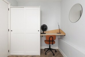 Une pièce avec une porte blanche, un bureau avec une chaise, un ventilateur fixé au mur et un miroir rond.