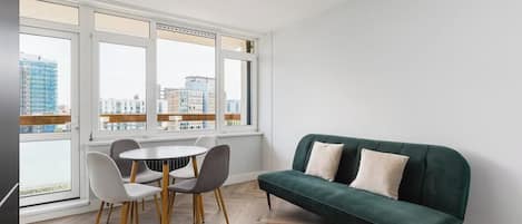 A modern room with a dining table, chairs, a green sofa, and large windows offering a city view.