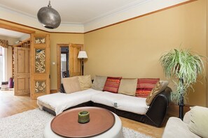 A living room with a sectional sofa, a round coffee table, a potted plant, and a wooden door with decorative panels. - New! Edwardian 4BR Gem | Garden and Free Parking (Greater London)