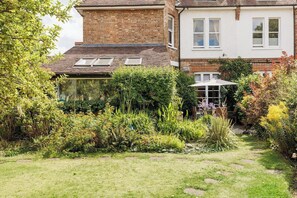 A residential property with a brick house, a garden with various plants, and a pathway leading to a conservatory. - New! Edwardian 4BR Gem | Garden and Free Parking (Greater London)