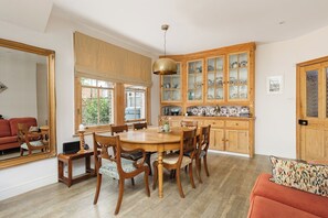 A dining area with a wooden table and chairs, a kitchen with wooden cabinets, and a large mirror on the wall. - New! Edwardian 4BR Gem | Garden and Free Parking (Greater London)